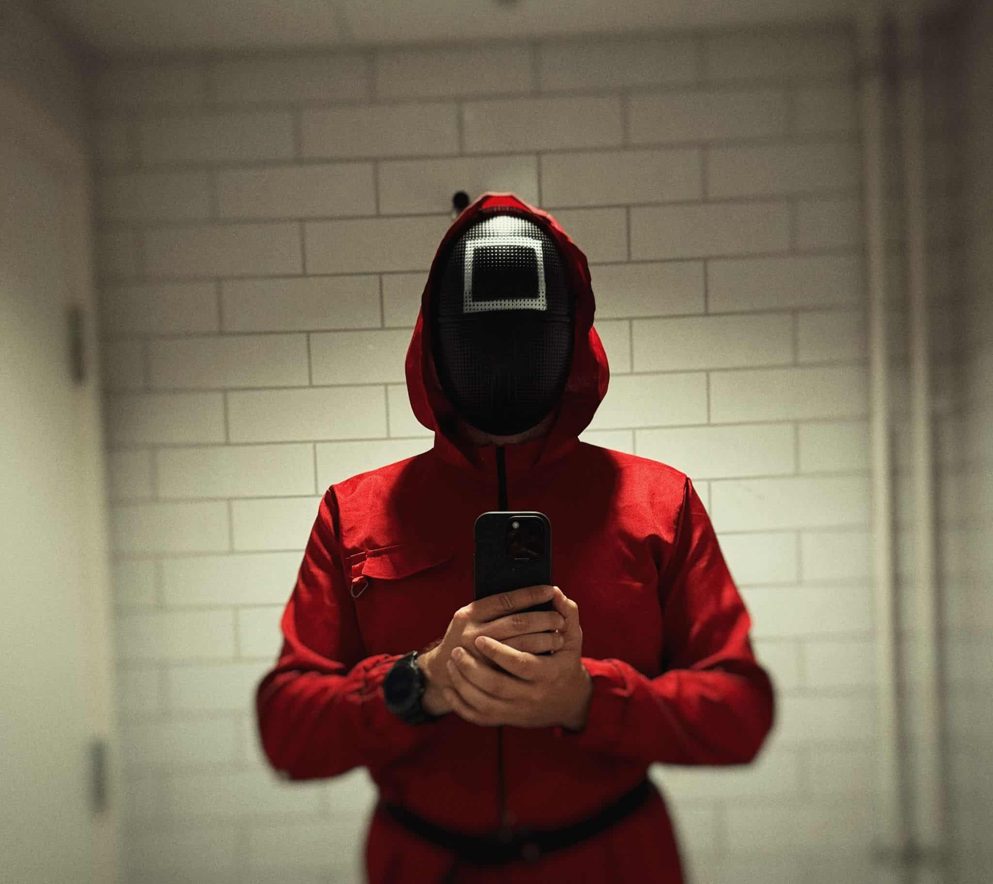 Person i rød Squid Game-dragt med firkantmaske holder en telefon.