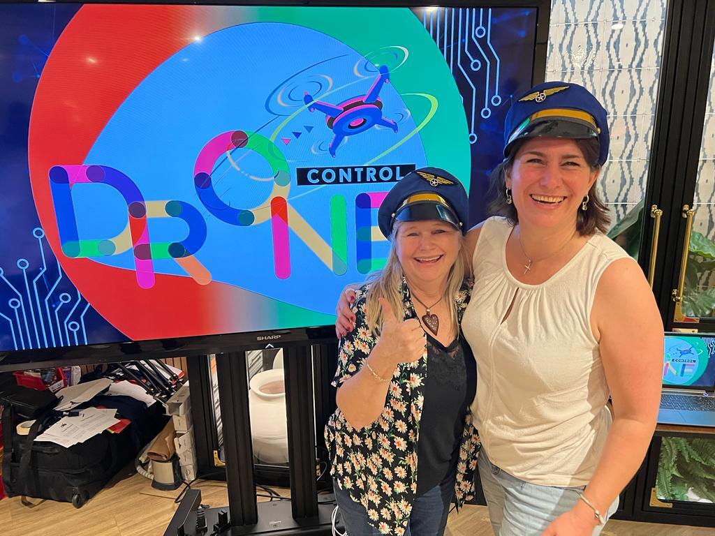 To glade kvinder med pilotkasketter foran en skærm med "Drone Control" logo.